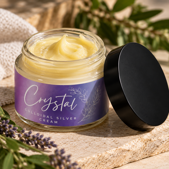 Open glass jar of pale yellow cream with a purple label reading Crystal Colloidal Silver Cream, black lid beside it, on stone with lavender sprigs.