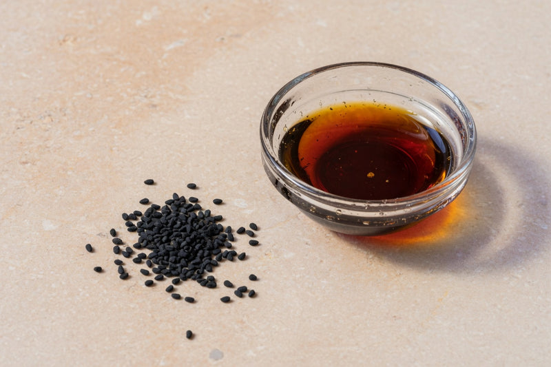 Tiny black nigella seeds beside a glass dish of dark amber oil.