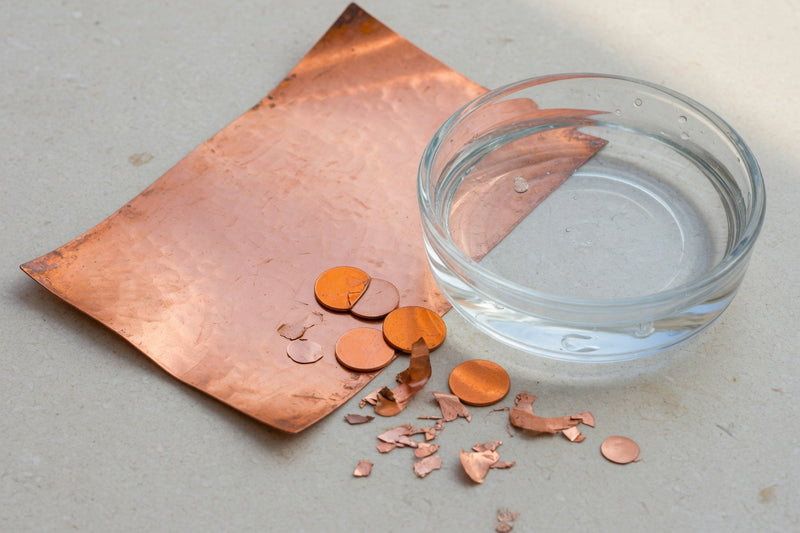 Warm copper metal beside a glass dish of clear water on pale limestone.