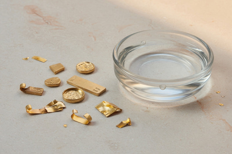 Yellow gold metal beside a glass dish of clear water on pale limestone.