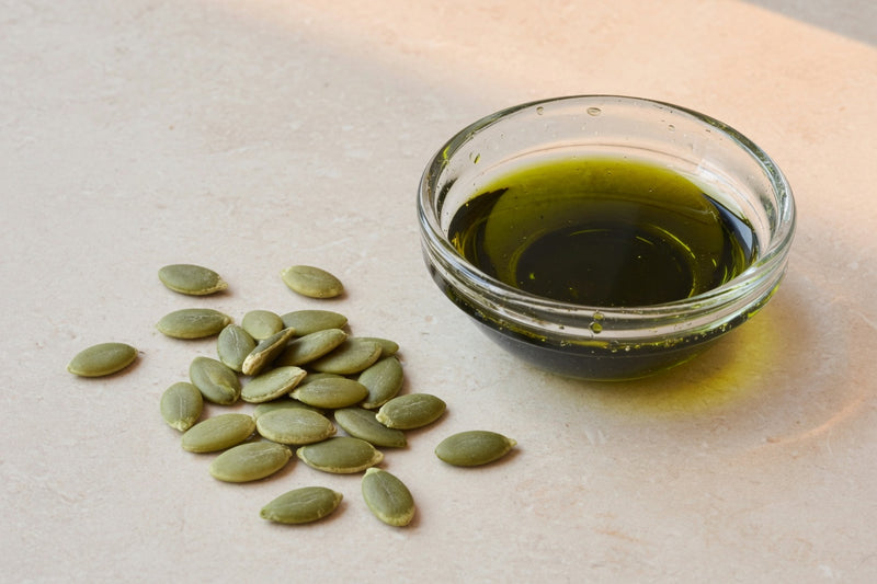 Green pumpkin seeds beside a glass dish of dark green oil.